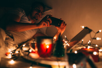 Yoiung man chilling out at home on Christmas eve, using his mobile phone. Beautiful warm ambient,