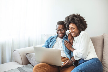 Young black couple sit on the sofa using laptop. Black couple using laptop at home look at each other. Overjoyed african American couple sit relax on cozy couch look at laptop screen triumph winning  - Powered by Adobe