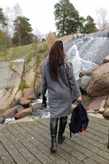 Fototapeta premium A young woman in a coat with a backpack in her hand looks at the waterfall. Kotka town park, Finland.