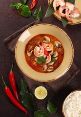 Thai cuisine. Tom yum. Coconut milk soup with shrimp, mushrooms, tomatoes, galangal root, lemongrass, chili pepper in a beige bowl on a brown table. Rice, lime. Background image, copy space, top view