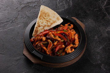 Mexican cuisine. Fajitas. Chicken fillet, fried sweet pepper, beans and tomato sauce. Tortilla a black iron pan on black table. Background image, copy space. Restaurant menu