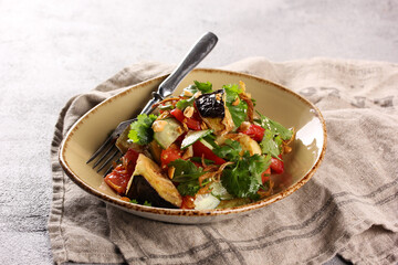 Oriental cuisine. Mr. Chaw's salad. Salad from fried eggplant, tomatoes, cucumbers, chili peppers, onions and fresh herbs with sauce in a beige plate on a grey table. Background image, copy space