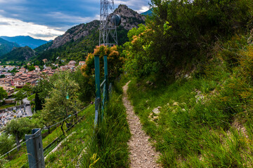 A picturesque view of a hiking path in the French Alps mountains on a cloudy summer evening next to the panoramic view of a French village (Puget-Theniers, Alpes-Maritimes, Provence, France)