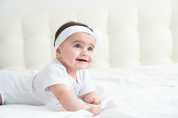 Cute healthy baby girl 6 months smiling in a white bodysuit lying on a bed on white bedding.