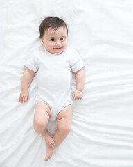 funny healthy baby girl 6 months in white bodysuit lying on bed on white bedding. top view.