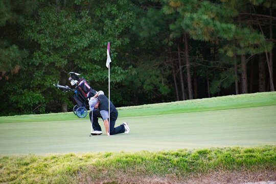 An Adult Man Golfer Picking Up A Golf Ball On The Green With A Pin On A Hard And Difficult Luxury Country Club Golf Course With A Golf Bag And Clubs Behind Him