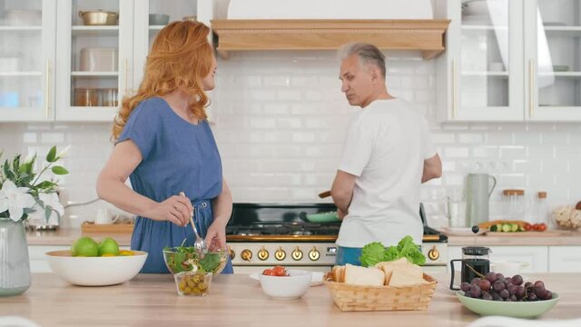 Medium shot of pretty middle-aged housewife standing at kitchen table, making vegetable salad and talking to grey-haired man frying eggs on background