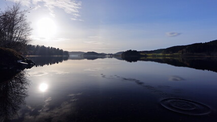 Wide panorama of lake with half solar halo in the blue sky and the reflection as a mirror image in the water. The continue ripples on the still water surface. Beautiful Scandinavia scenery.