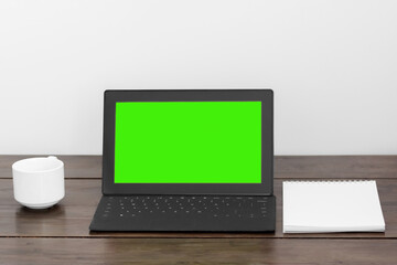 Fototapeta premium A tablet with a green screen on a wooden table, a white cup and notepad stands near.