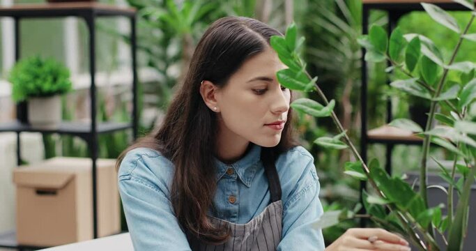 Close up of happy beautiful Caucasian woman entrepreneur in good mood checking plant and taking notes in floral center. Joyful female florist at work in flower store writing in planner. Job concept