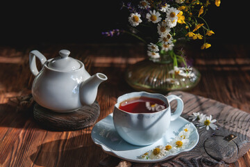 Cup of tea with chamomile flowers and a white teapot on a rustic wooden background. Rustic still life, authentic setting