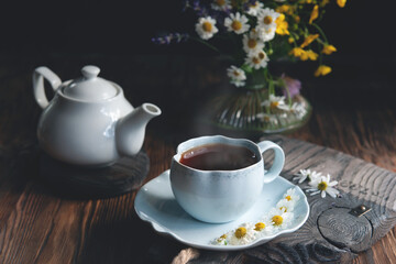 Cup of tea with chamomile flowers and a white teapot on a rustic wooden background. Rustic still life, authentic setting