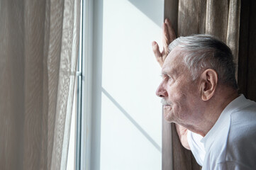 Fototapeta premium gray-haired old man, forced to remain in quarantine due to the covid-19 coronavirus, looks out the window with a disconsolate expression. Loneliness of old people. Stay home in the pandemic