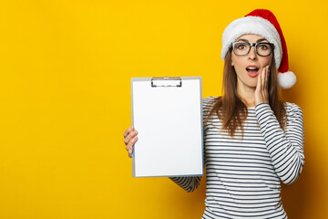 Young woman with a surprised face in a Christmas hat holds a clipboard on a yellow background. Banner. Copy space