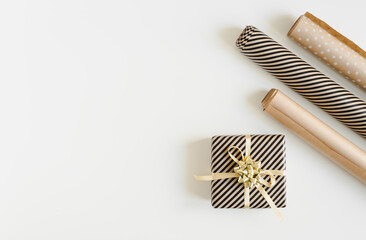 Christmas gift box wrapped in paper with golden ribbon and three kraft paper rolls on white background, top view, copy space. DIY gift wrapping idea flat lay
