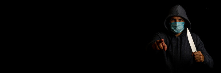 Young man in a medical mask points a finger while holding a knife on a black background. Banner.