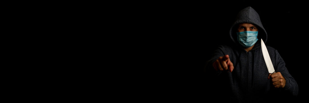 Young man in a medical mask points a finger while holding a knife on a black background. Banner.