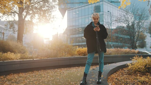 Young caucasian short haired blond woman using phone in the city park in front of office building in autumn. High quality 4k footage
