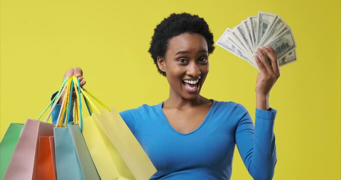 Delighted african american woman holding dollar banknotes and showing her shopping bags
