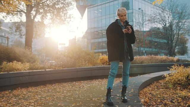 Young caucasian woman using phone in the city park in front of office building in autumn. High quality 4k footage