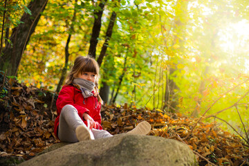 Little child girl with red jacket, sitting on a stone in autumn forest scenery with copy space and sunflare. Varese, Italy.