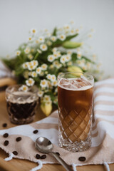Black ice coffee with milk in the vintage glass on the ligh background, wooden tray and beige striped tablecloth, coffee beans, bouquet in the background