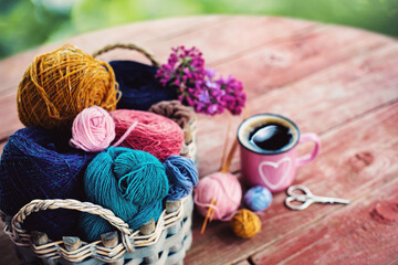 Fototapeta na wymiar Women's hobby. Crochet and knitting. Working space. Multicolored skeins in the basket, needles, scissors and pink mug with tea on the wooden table in the garden. 