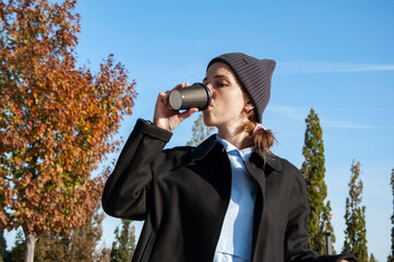 a girl wearing a hat drinks coffee from a takeaway cup in autumn