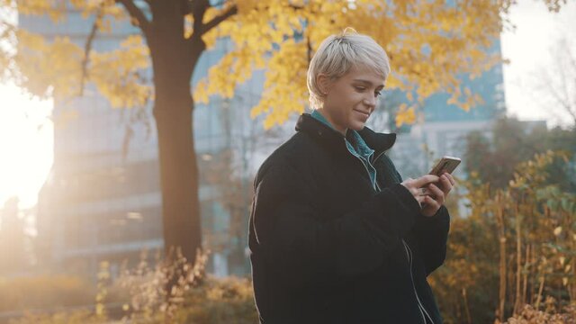 Young caucasian blond woman using phone in the city park in front of office building in autumn. High quality 4k footage
