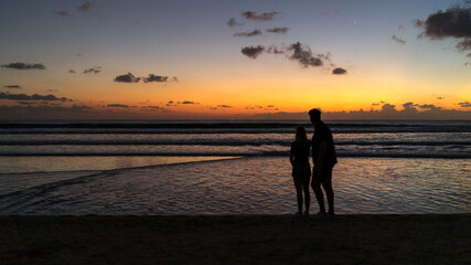 A silhouette of a couple on the beach watching the golden sunset in Kuta, Bali. Soft focus, film noise