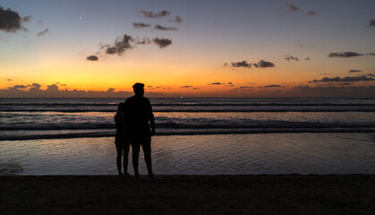 A silhouette of a couple on the beach watching the golden sunset in Kuta, Bali. Soft focus, film noise