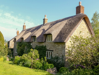 Quintessential old thatched cottage built in local stone, in sunshine. Rural setting with natural shadows under a predominantly blue sky. Pretty cottage garden. England. - 390496197