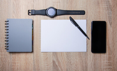 Smartphone and smartwatch next to a notebook and blank card  on a wooden desk. There is also pen on...