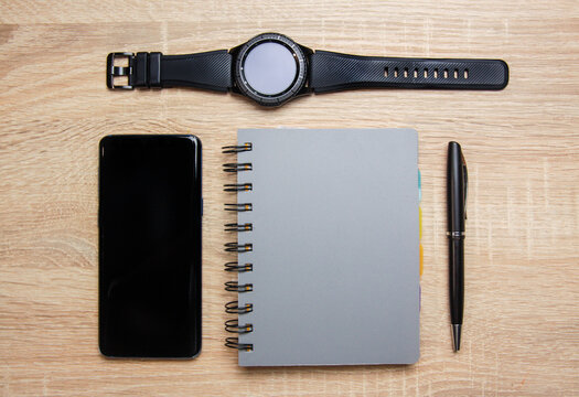 Smartphone and smartwatch next to a notebook on a wooden desk. There is also pen on the desk.