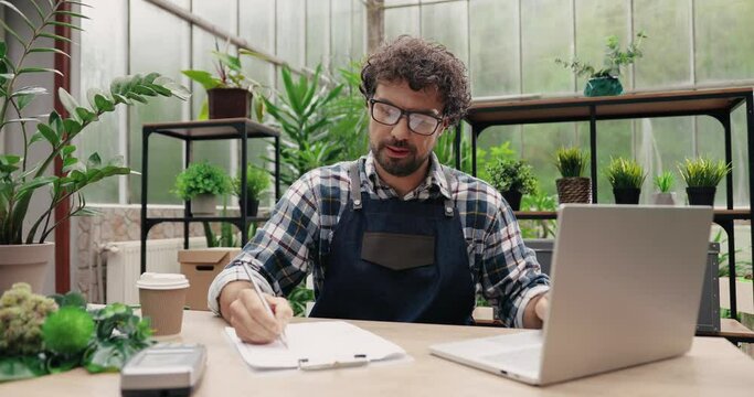 Portrait of Caucasian man entrepreneur in glasses sitting in own flower shop, typing on laptop and writing down in planner. Young male employee in floral store working on computer. Florist concept