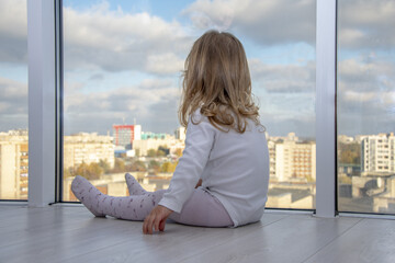 A little girl sits on the floor of an apartment in front of huge Windows and looks down at the city. Concept: children's loneliness, dreaminess and observation.
