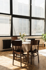 Bright living dinning room interior design. Windows, round dinner table, chairs, flowers bouquet on linen tablecloth. Warm sunlight shadows on the wooden parquet. Retro vintage bohemian home concept.