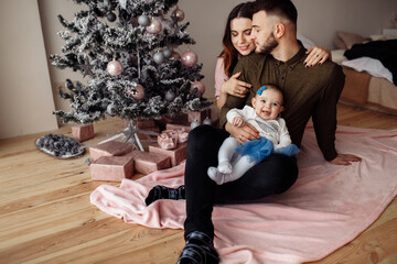 Happy family with little daughter sit near christmas tree, caring dad hold cute toddler in arms, beautiful wife hug charming husband, enjoy tender moments, holidays at home concept