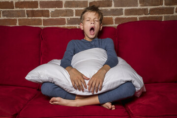 Tired Youth Boy In Pajamas With Pillow Sitting On Couch