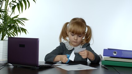 Child schoolgirl learns lessons at home sitting at table cutting with scissors shapes out of paper. Kid girl distance education. Studying during coronavirus quarantine. White background