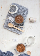 Two chocolate mousse jars on a wooden table. One on a knitted cloth and the other next to a blue napkin and several spoons.