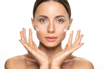 Obraz na płótnie Canvas Young woman with flawless skin applying nourishing cream on her face cheek and fingers, looking at camera, studio white background. Girl facial treatment. Skin care, beauty cosmetic concept