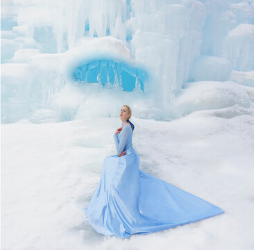 Winter Fairy Tale On An Ice Waterfall A Beautiful Girl With Long Blonde Hair And A Blue Dress With A Long Train.Snow Queen Luxurious Lush Full Dress Waving Silk Fabric Skirt Long Train. 