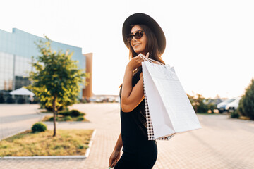 happy attractive elegant girl, in a black dress and hat, wearing dark glasses, holding shopping...