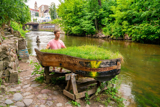 Young man playing abandoned gran piano covered with grass, rundown piano by a river bank Vilnia river in Uzupis artists quarter in Vilnius