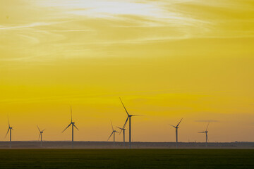 many windmills stand on a large field spinning in the evening against a sunset orange sky....