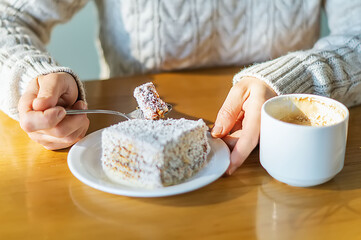 A young woman is sitting at a cafe table in a warm knitted sweater and using a teaspoon to cut a piece of delicious cake. Cup of coffee on the table. Close-up, selective focus