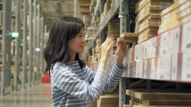 Asian woman working or shopping on shelves at large warehouse retail store industry. Rack of furniture and home accessories store. Interior of cargo in ecommerce and logistic concept. Lifestyle
