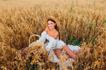 Young beautiful woman with straw hat sitting at golden oat field near the basket with ears of oats.
