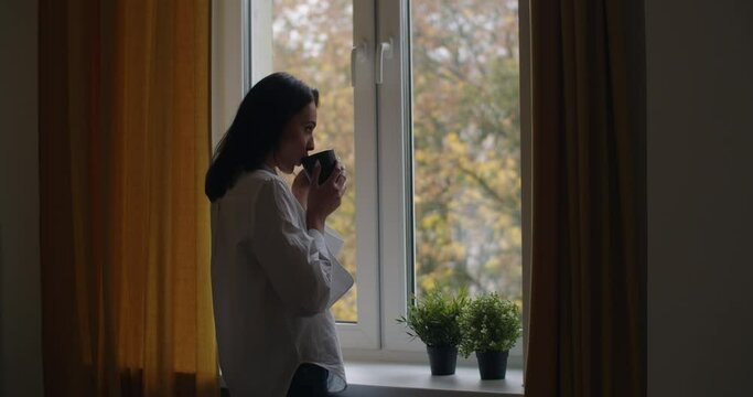 Side portrait of pensive girl standing by window with cup of tea slow motion. Young woman drinking hot coffee looking outside at autumn landscape copy space. Loneliness depression solitude concept
